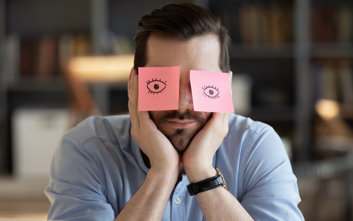 Bărbat doarme la birou cu două post-it-uri pe care sunt desenați ochi deschiși