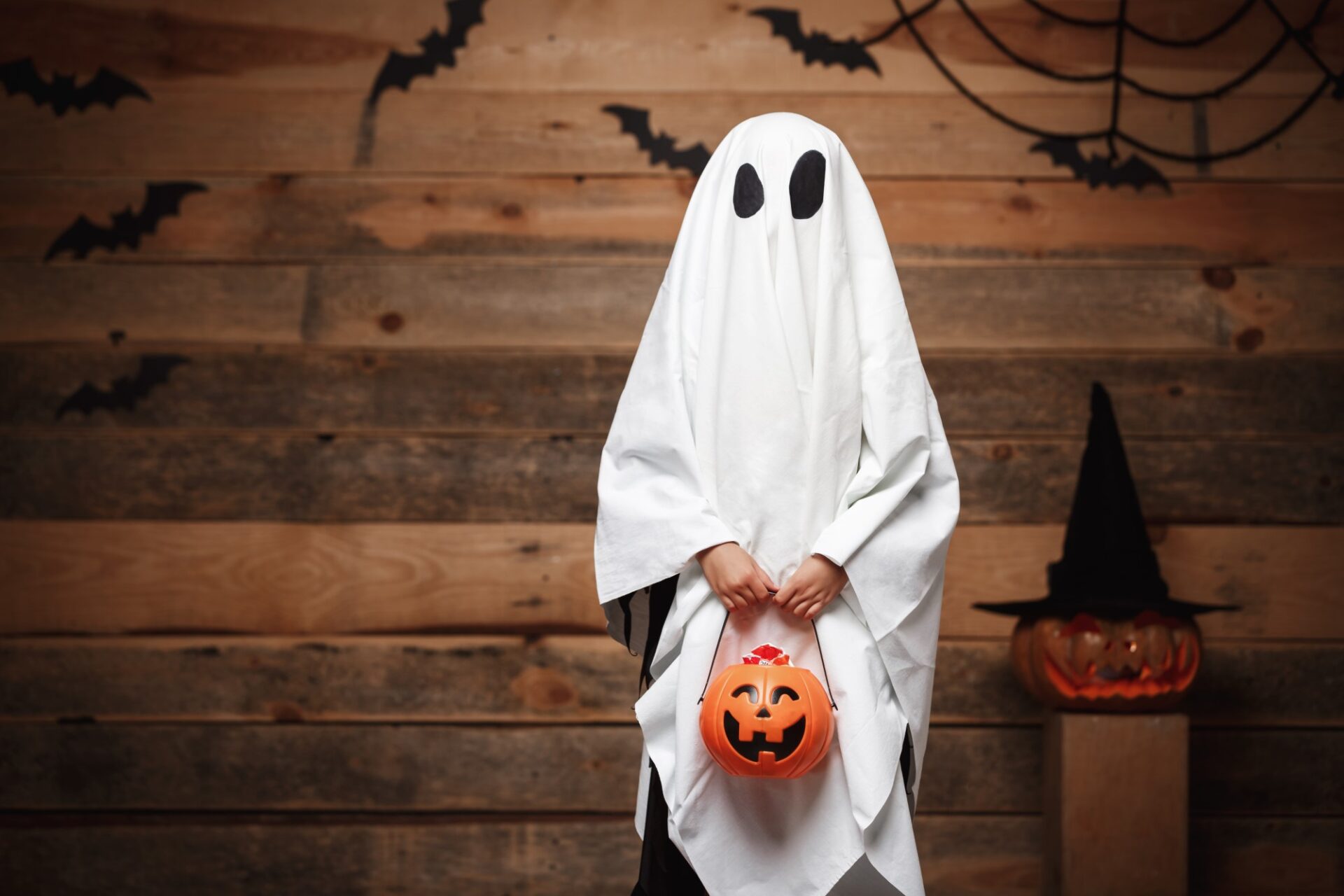 copil costumat in fantomă de Halloween, cu dovleac în mână, lilieci pe pereți