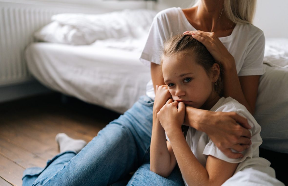 Mama îmbrățișează strâns fiica tristă, stând pe jos, lângă pat
