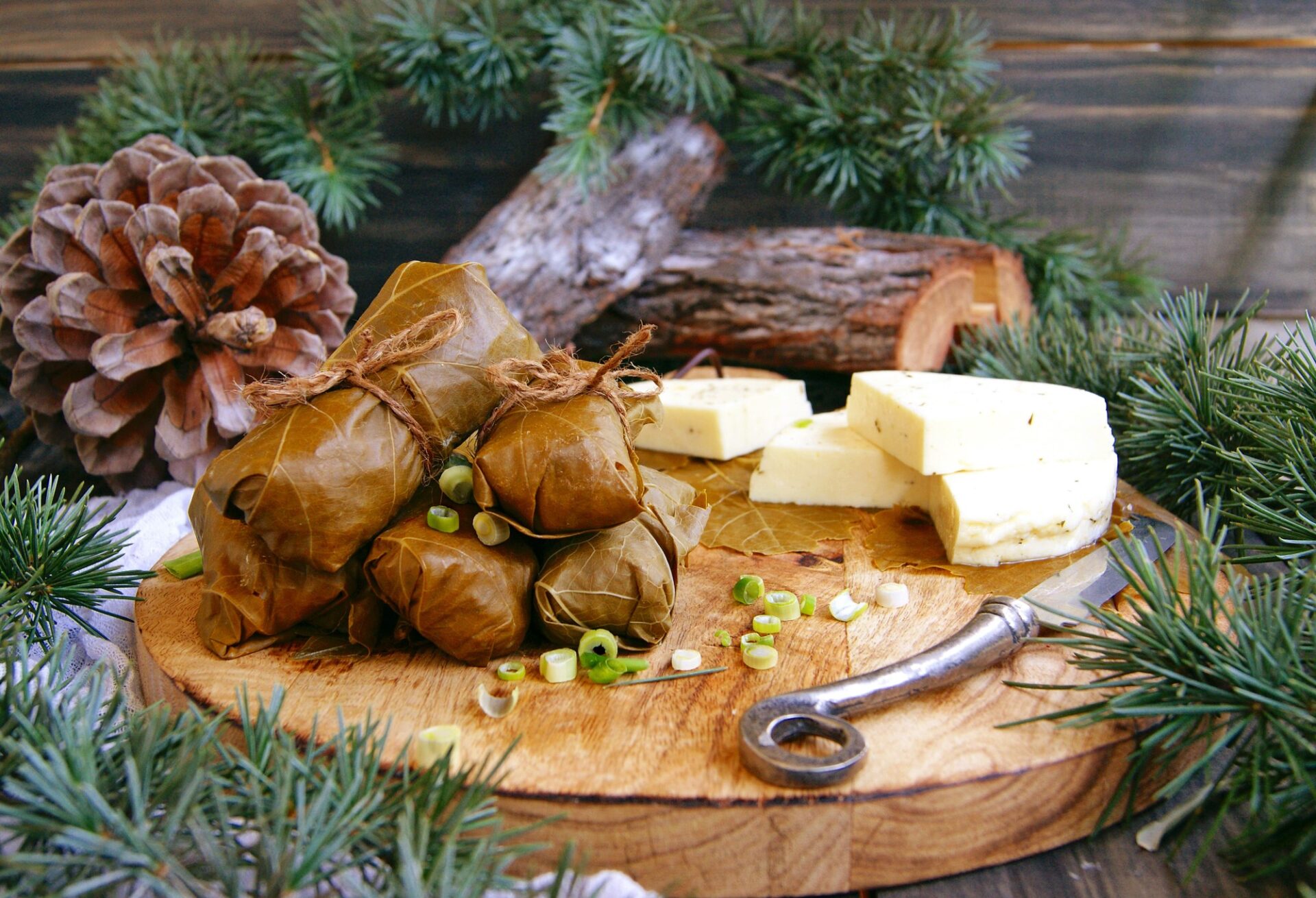 sarmale în foi de viță gourmet, legate cu sfoară și pe un platou cu crengi de brad, conuri și brânză gourmet