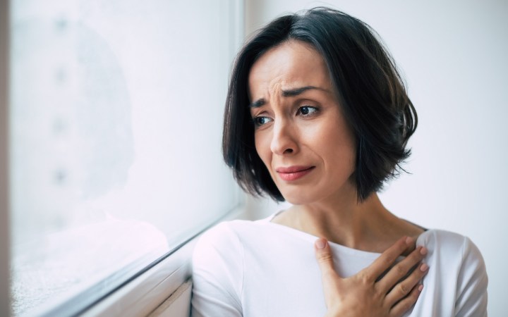 femeie tânără se uită pe geam și plânge cu o mână pe piept