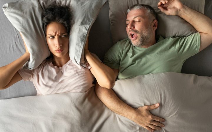 cuplu în pat, bărbatul sforăie și femeia își acoperă urechile cu perna